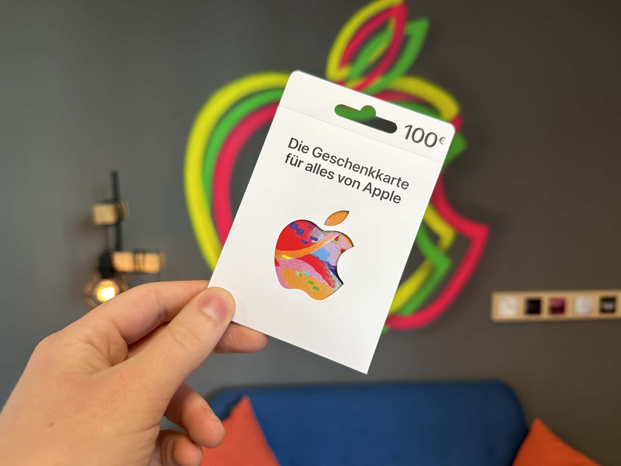 100 Euro Apple Gift Card in der Hand vor Apple Logo Wand